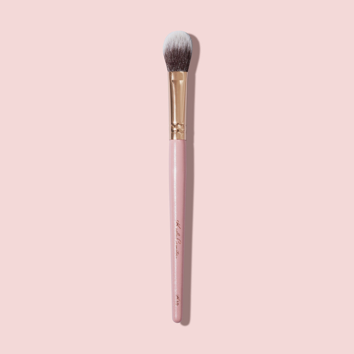 Der K10 Highlighter Brush mit rosa Griff und Borsten mit weißen Spitzen auf einem hellrosa Hintergrund.