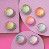 Acht Tiegel der Opal Moonstone Loose Multichrome Eyeshadows auf pastellfarbenen geometrischen Formen.