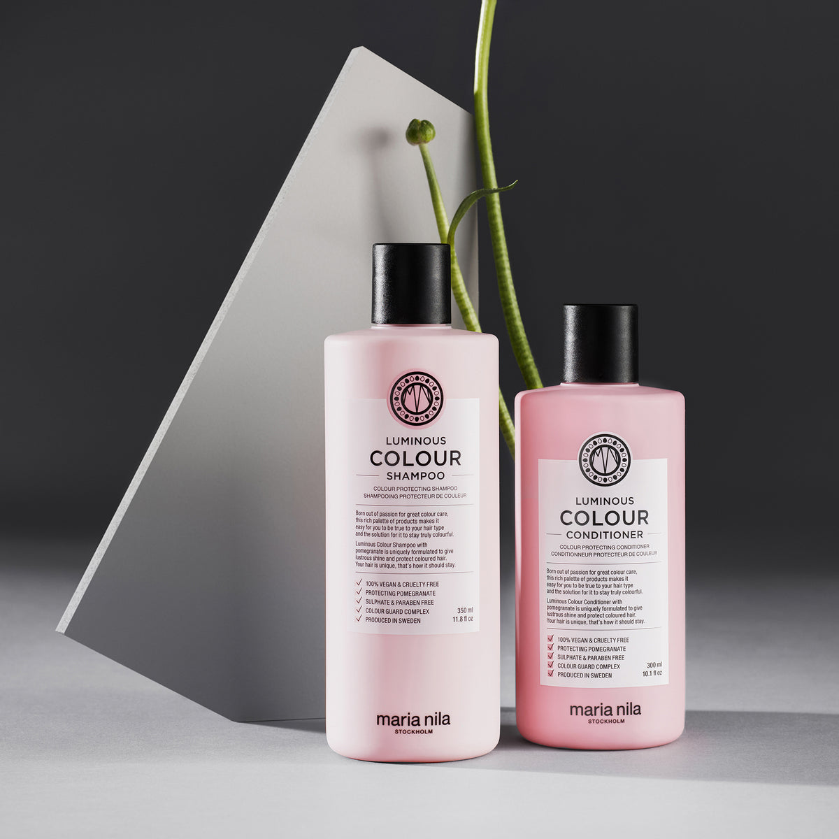 Zwei rosafarbene Flaschen von maria nila Luminous Colour Conditioner und Shampoo sind vor einem grauen Hintergrund platziert.
