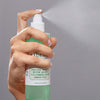 Hand sprüht das Mario Badescu Facial Spray mit Aloe, Gurke und grünem Tee aus einer grünen Flasche .