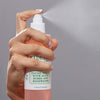 Hand sprüht das Mario Badescu Facial Spray mit Aloe, Kräutern und Rosenwasser vor grauem Hintergrund.