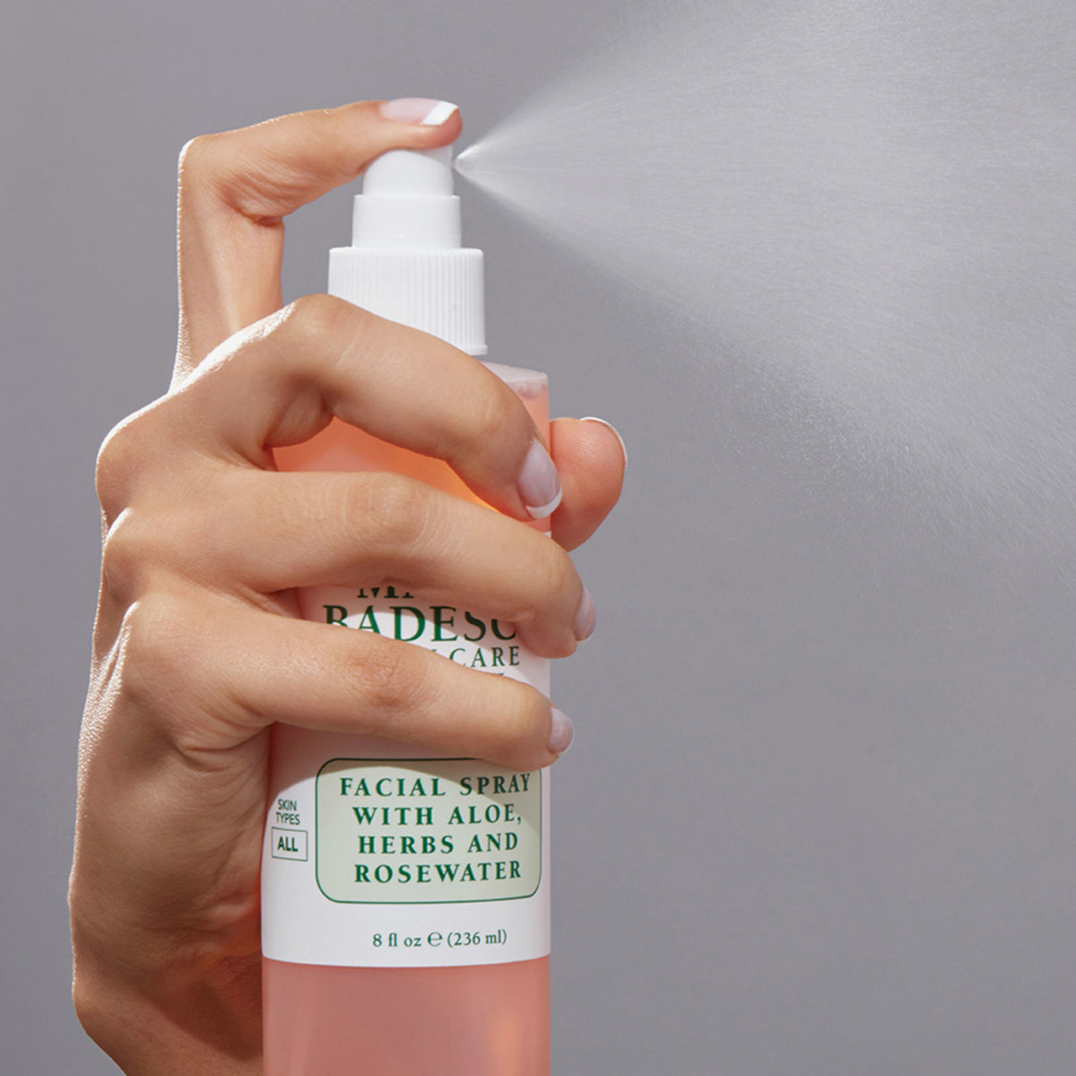 Hand sprüht das Mario Badescu Facial Spray mit Aloe, Kräutern und Rosenwasser vor grauem Hintergrund.