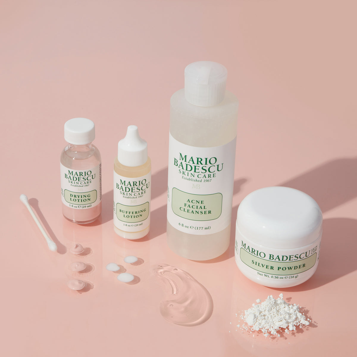 Mario Badescu Acne Facial Cleanser, drei weitere Hautpflegeprodukte, Wattestäbchen und Proben auf einem rosa Hintergrund.