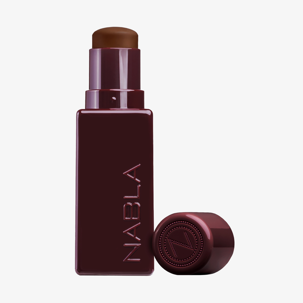 Eine burgunderfarbene Tube des NABLA Cosmetics Art Of Skin Foundation Stick mit abgenommenem Deckel, der das braune Produkt im Inneren zeigt.