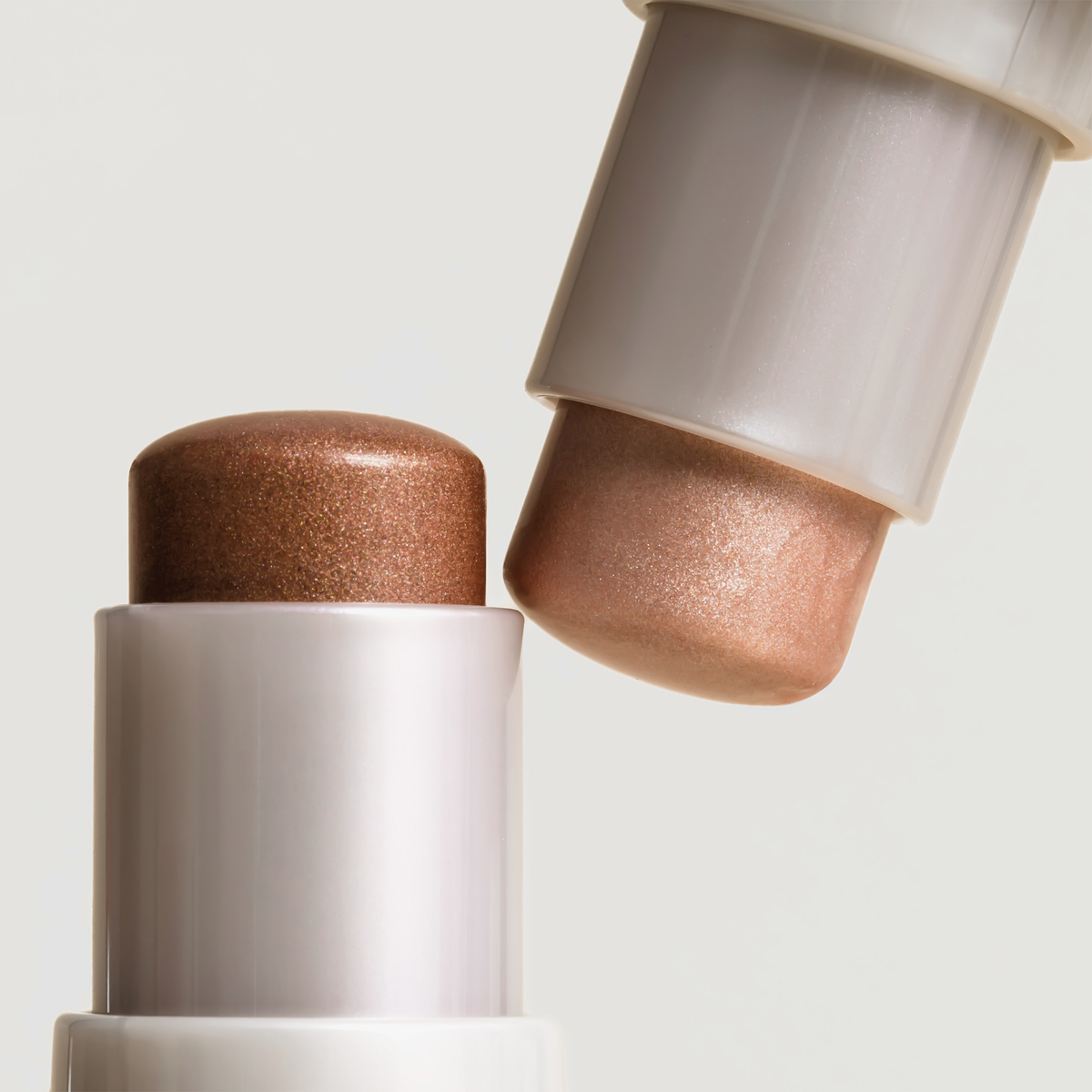 Zwei „Art Of Skin Glazing Sticks“ von NABLA Cosmetics in den Farben Bronze und Champagner, abgebildet auf hellgrauem Hintergrund.