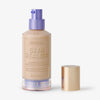 Eine Flasche Skin Realist Tinted Balm in der Farbe 1.5 Light mit einem violetten Pumpspender und einem goldenen Deckel daneben.