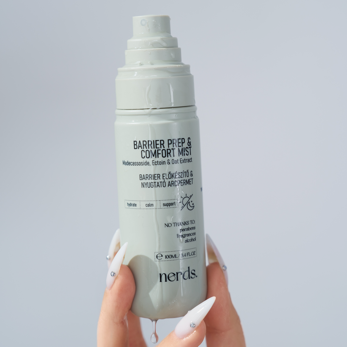 Eine Hand mit gepflegten Nägeln hält eine graue Flasche „NERDS. Barrier Prep & Comfort Mist“ in der Hand.