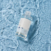 Eine durchsichtige Flasche des ROUND LAB 1025 Dokdo Toners mit weißem Etikett schwimmt auf dem plätschernden blauen Wasser und bringt so ihr minimalistisches Design zur Geltung.