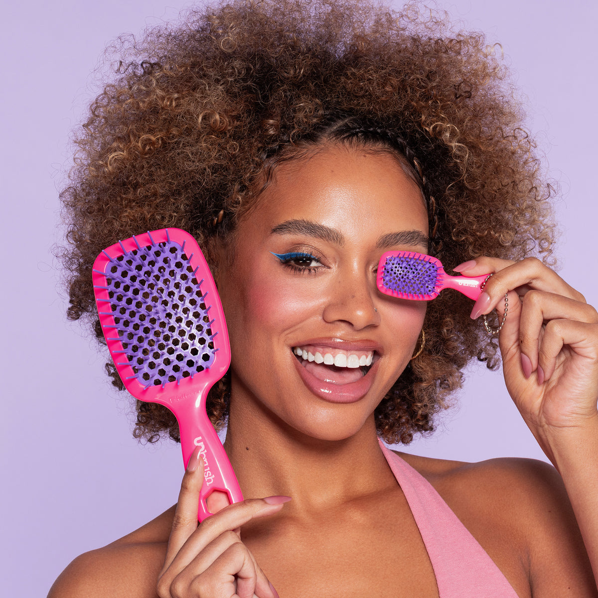 Frau mit lockigem Haar lächelt und hält eine große sowie eine kleine pink-lila UNbrush® Haarbürste im Keychain-Design hoch.