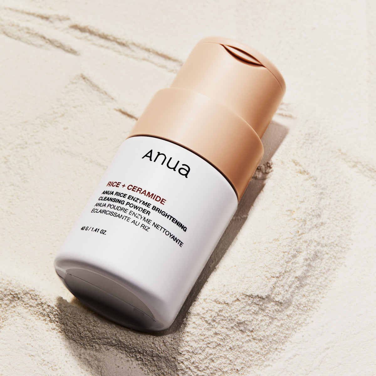 Eine Flasche Rice Enzyme Brightening Cleansing Powder liegt auf einer strukturierten, sandigen Oberfläche.
