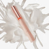 Das Colour Cushion Juicy Lip Oil mit rosafarbener Tube und roségoldener Kappe ist auf einem weiß gespritzten Flüssigkeitsuntergrund abgebildet.