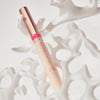 Ein Catrice Colour Cushion Juicy Lip Oil in Rosé und Pink liegt dekorativ in weißem, organisch geformtem Dekor.