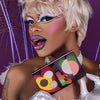 Eine Person mit buntem Make-up und Perücke hält die leuchtende Love is Love-Palette vor einem violetten Hintergrund.