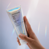 Eine Hand hält eine Tube Living Proof. High-Shine Gloss vor einem weichen, schillernden Hintergrund.