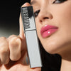 Eine Person mit glänzenden rosa Lippen hält eine silbern glänzende Tube LIP PHENOM™ von Mesauda Beauty an ihr Gesicht.