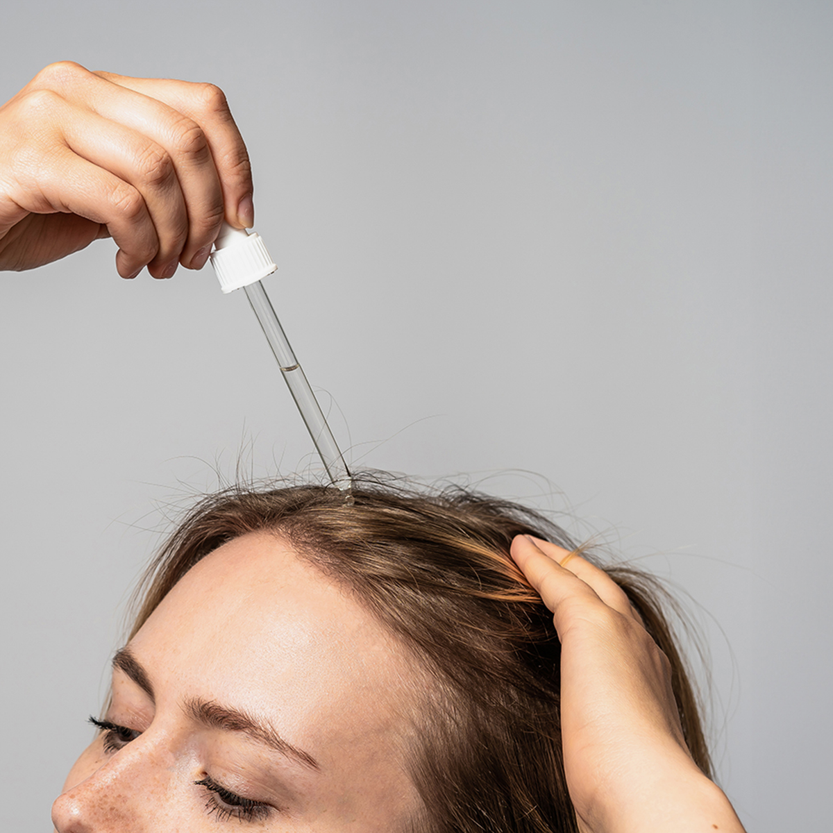 Eine Frau trägt Bio-Pilixin® Serum+ Women von Scandinavian Biolabs mit einer Pipette auf ihre Kopfhaut auf, vor einem schlichten grauen Hintergrund.