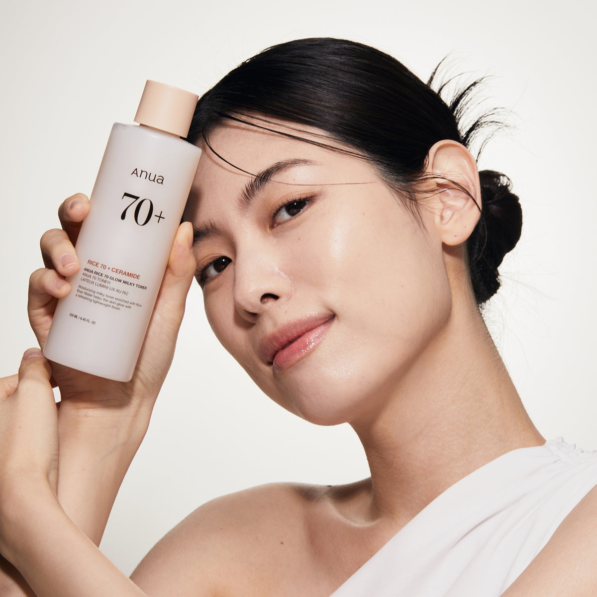 Lächelnde Frau, die vor einem hellen Hintergrund eine Flasche Rice 70 Glow Milky Toner nah an ihr Gesicht hält.