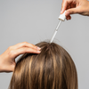 Eine Frau scheitelt ihr braunes Haar und trägt Bio-Pilixin® Serum+ Women von Scandinavian Biolabs mit einer Pipette auf ihre Kopfhaut auf.