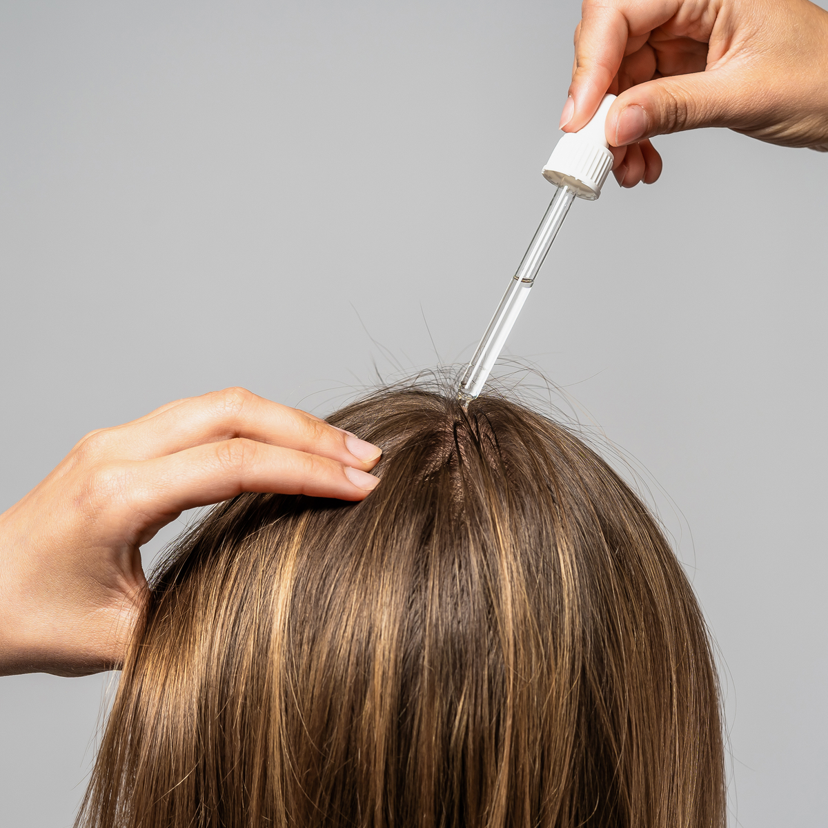 Eine Frau scheitelt ihr braunes Haar und trägt Bio-Pilixin® Serum+ Women von Scandinavian Biolabs mit einer Pipette auf ihre Kopfhaut auf.