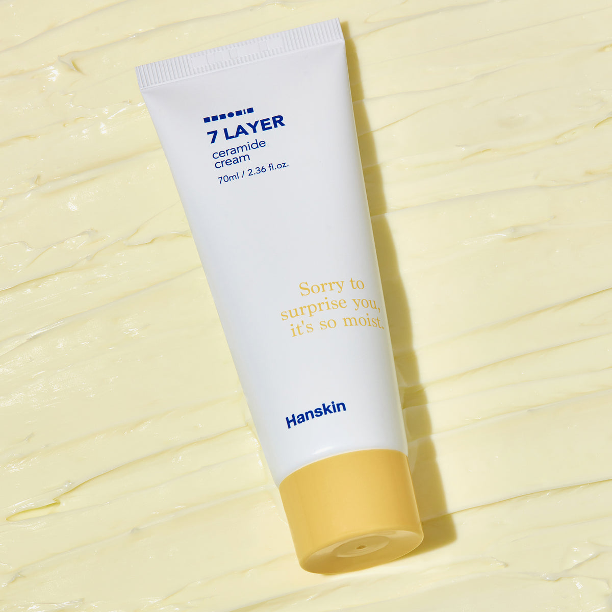 Weiße Tube Seven Layer Ceramide Cream auf einem cremegelben strukturierten Hintergrund.