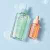 Zwei Flaschen maria nila Purifying Cleans und Head & Heal Soothing Serum ruhen auf einer hellblauen Fläche, umgeben von Tröpfchen.