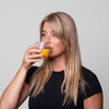Eine Frau mit langen blonden Haaren vor einem grauen Hintergrund, trinkt orangenes Bio-Silacumin von Scandinavian Biolabs aus einem Glas.