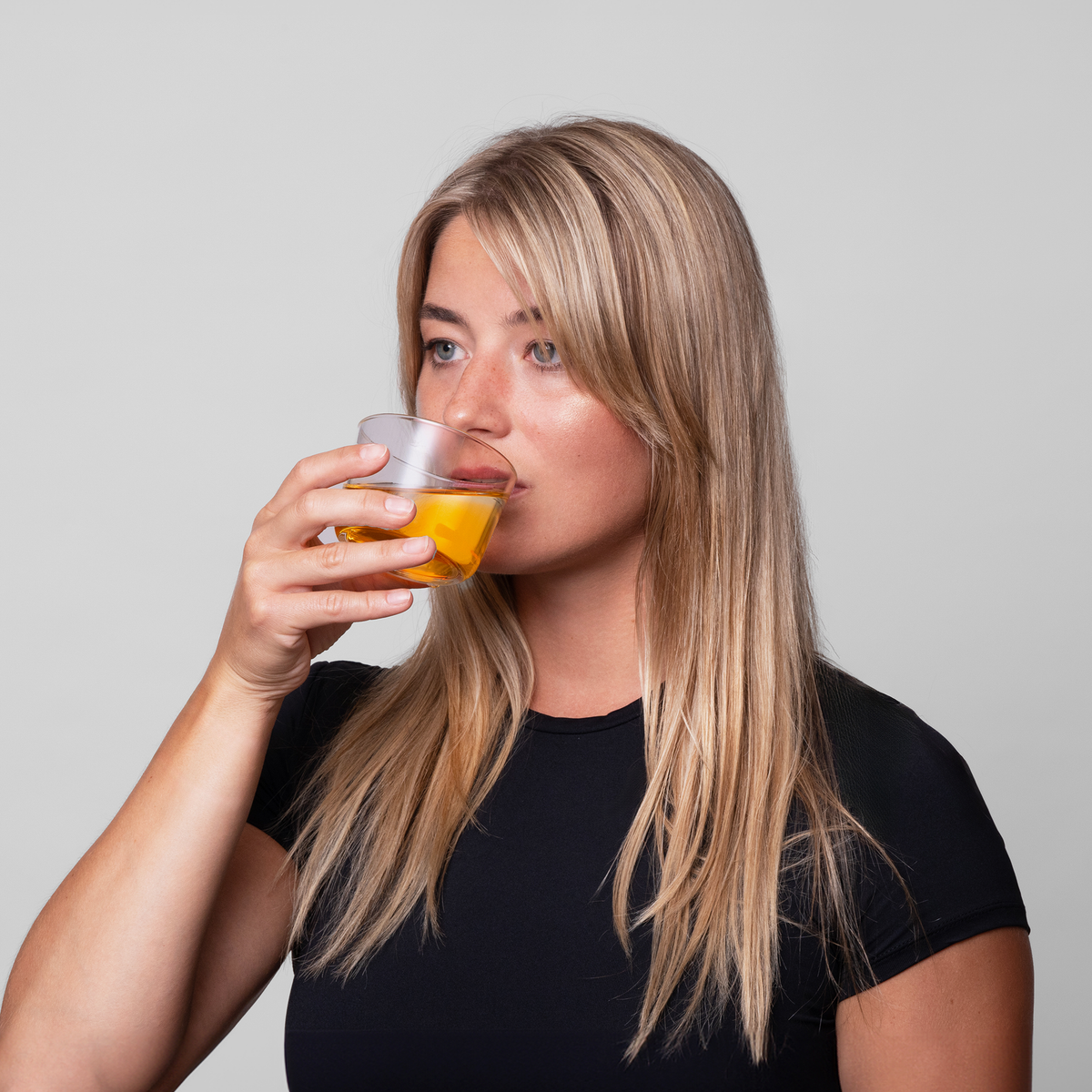 Eine Frau mit langen blonden Haaren vor einem grauen Hintergrund, trinkt orangenes Bio-Silacumin von Scandinavian Biolabs aus einem Glas.