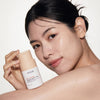 Lächelnde Frau mit strahlender Haut hält Rice Enzyme Brightening Cleansing Powder vor einem schlichten Hintergrund.