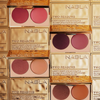 Make-up-Paletten von Two Reasons mit zwei Creme-Rouge-Tönen, die in einem Gittermuster angeordnet sind, wobei einige Paletten offen und andere geschlossen sind.
