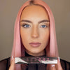 Eine Frau mit rosa Haaren demonstriert die Wirkung von Lash Out Mascara, indem sie ein Auge mit und eines ohne Mascara zeigt.