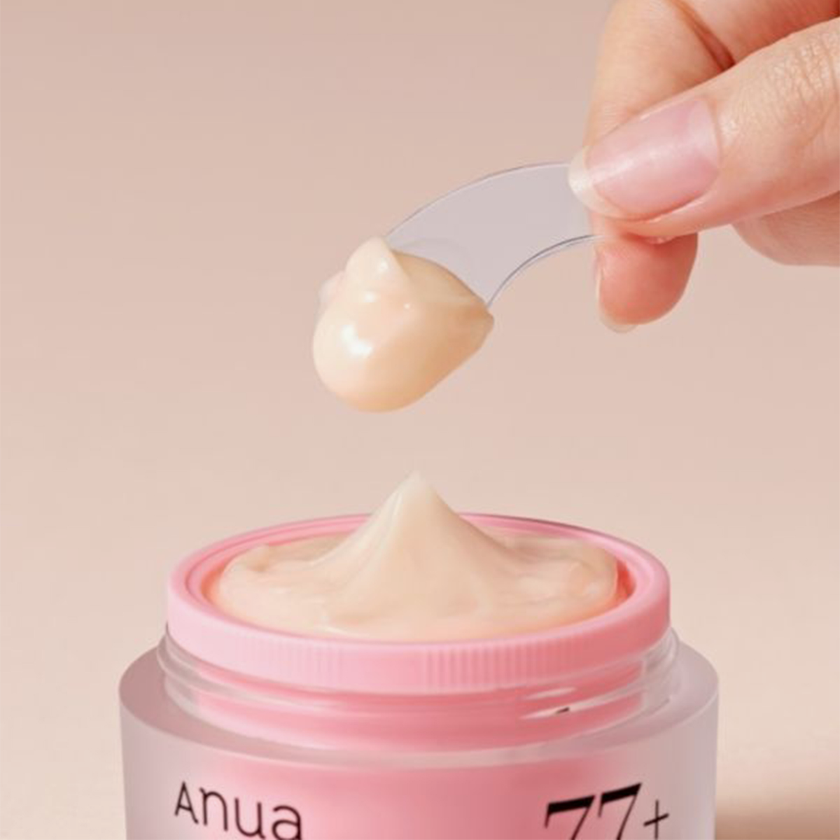 Eine Hand hält einen Spatel mit Creme über einem offenen, rosafarbenen Tiegel mit „ANUA Peach 77 Niacin Enriched Cream“.