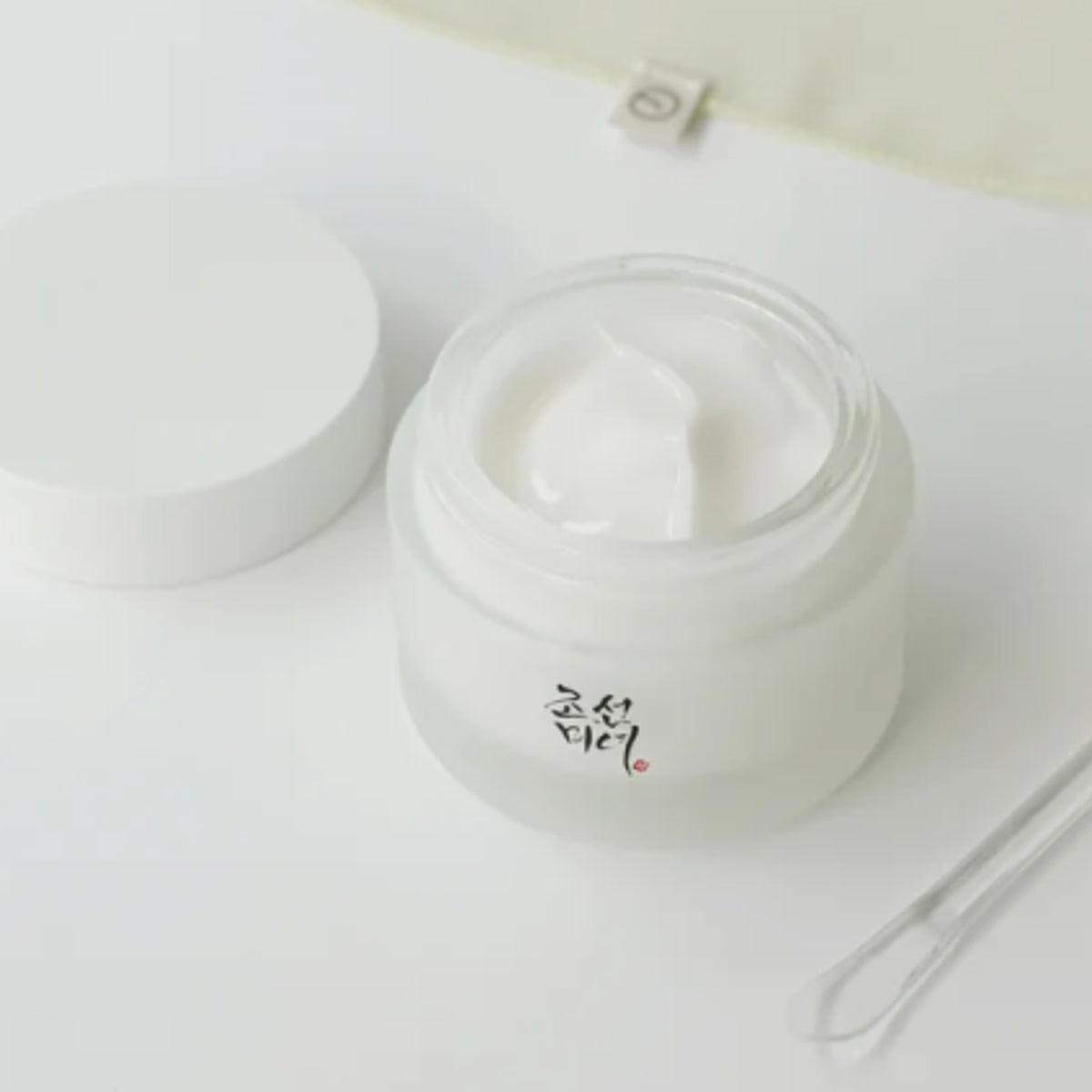 Ein offener Tiegel  Dynasty Cream auf einer weißen Oberfläche; daneben ein transparenter Spatel und der weiße Deckel