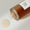 Nahaufname des Ginseng Essence Waters mit brauner Flüssigkeit und einem Tropfen auf beiger Oberfläche