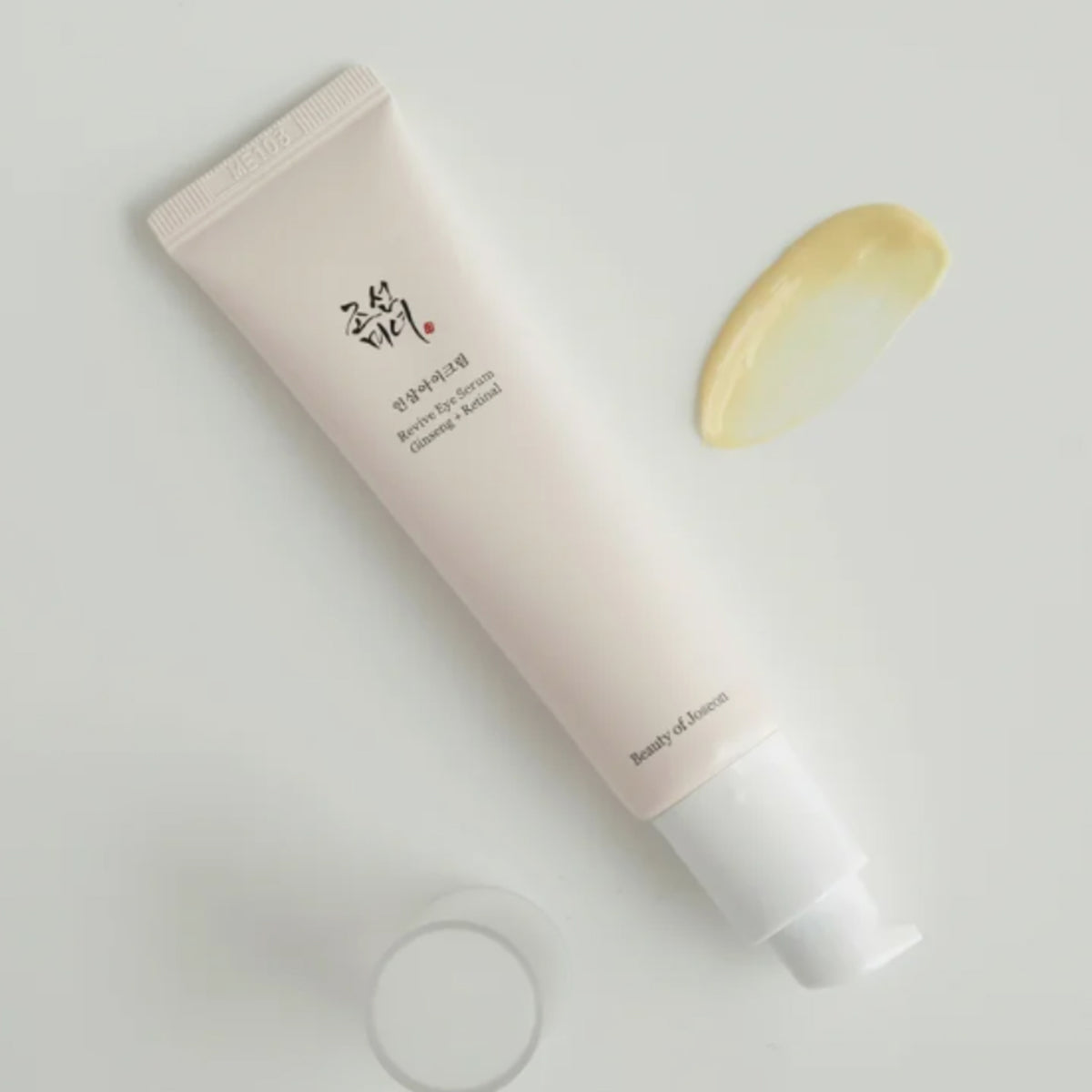Eine Pumptube Revive Eye Serum: Ginseng & Retinal von Beauty of Joseon ohne Deckel liegt neben gelbem Klecks Creme auf heller Oberfläche