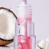 BLONDESISTER 3-IN-1 CUTE LIPPY LIP OIL mit rosa Tropfpipette, abgebildet neben aufgeschnittenen Kokosnussstücken.