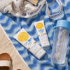 Auf dem Sand liegt ein Strandtuch mit einer SPF 50+ Everyday Body Lotion daneben eine blaue Wasserflasche und Sandalen.