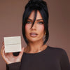 Model mit dunklem Haar und braunem Shirt hält die BPerfect Cosmetics x Katie Daley Perfect Powders Verapckung vor braunem Hintergrund.