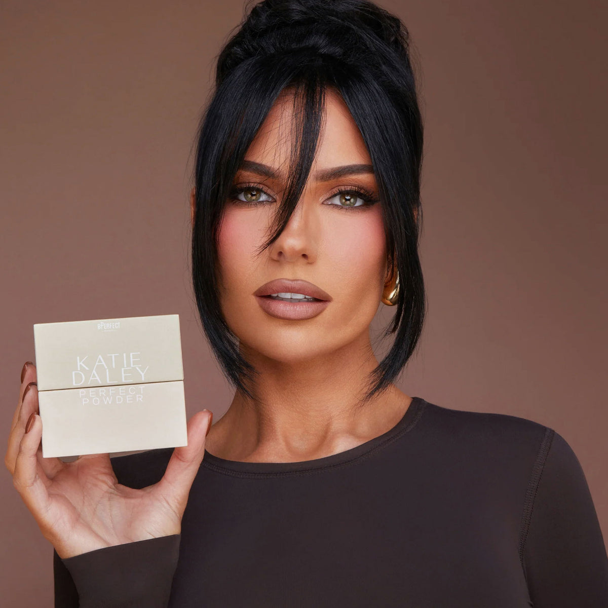 Model mit dunklem Haar und braunem Shirt hält die BPerfect Cosmetics x Katie Daley Perfect Powders Verapckung vor braunem Hintergrund.