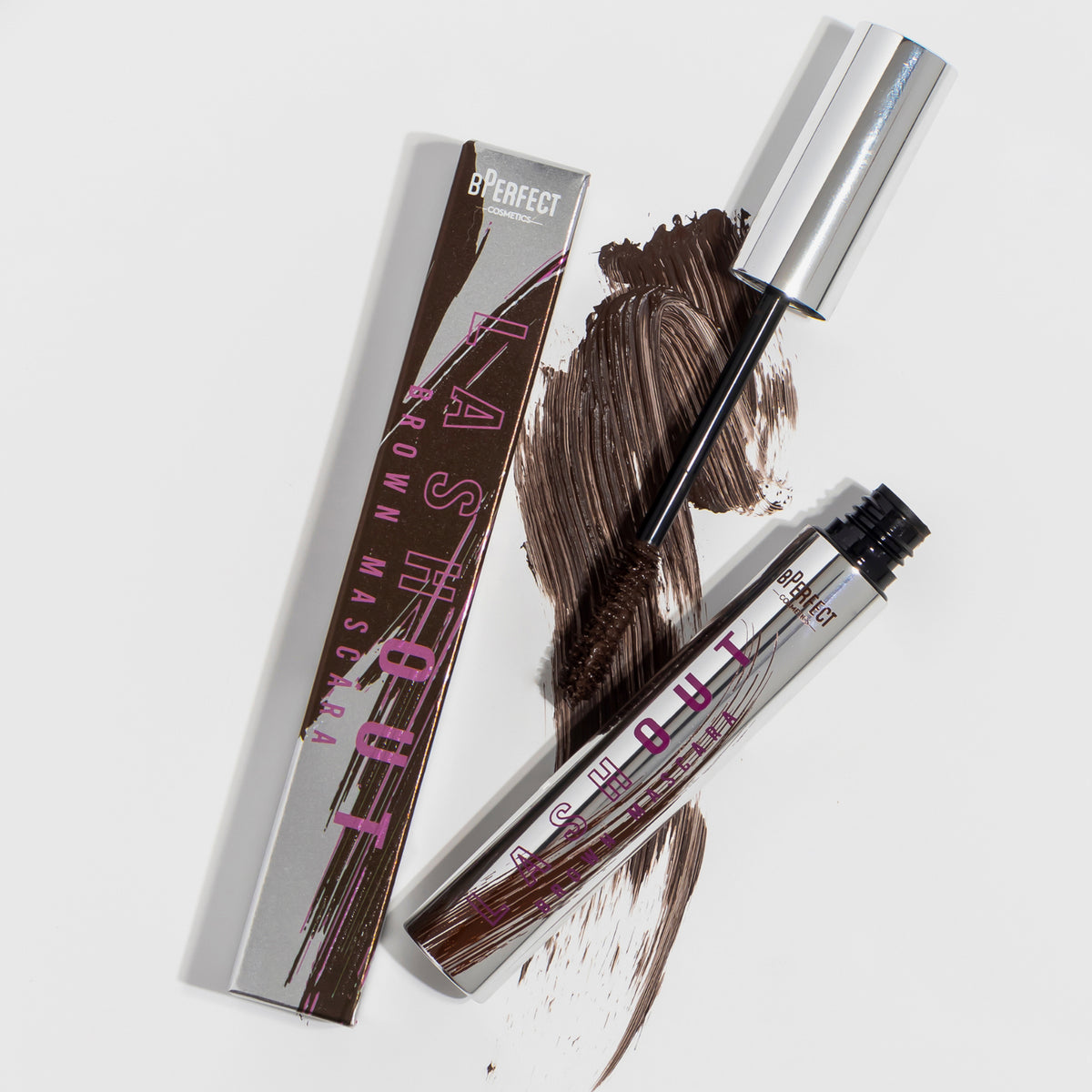 Die silberne Lash Out Mascara Brown von BPerfect liegt neben ihrer Schachtel, auf deren weißer Oberfläche braune Mascara verschmiert ist.