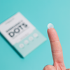 Ein durchscheinendes, rundes Emergency Dots with Tea Tree Oil-Pflaster, das auf einer Fingerspitze vor der Breakout+Aid-Verpackung angebracht ist.