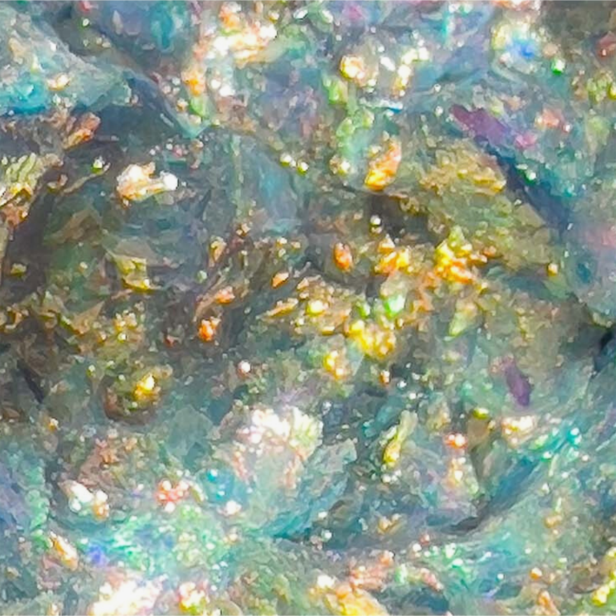 Nahaufnahme von Danessa Myricks Beauty Infinite Chrome Flakes Firefly, die eine farbenfrohe, schillernde Oberfläche mit blauen, grünen und golden schimmernden Flecken zeigen.