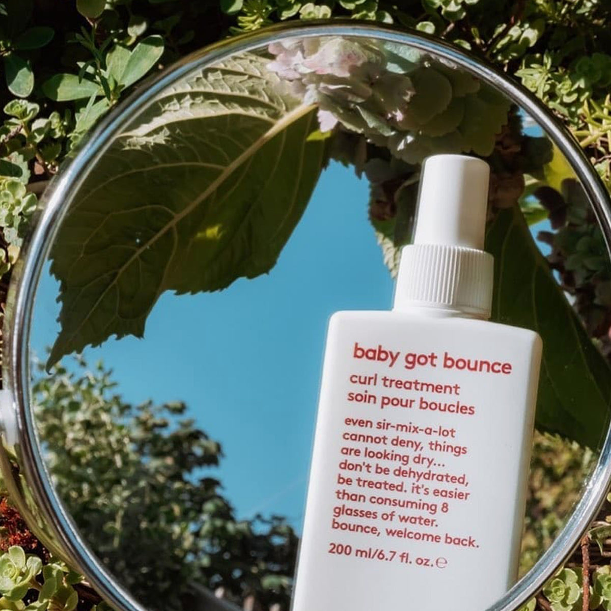 Eine Flasche Baby Got Bounce Curl Treatment spiegelt sich in einem Spiegel im Freien, umgeben von grünen Pflanzen.