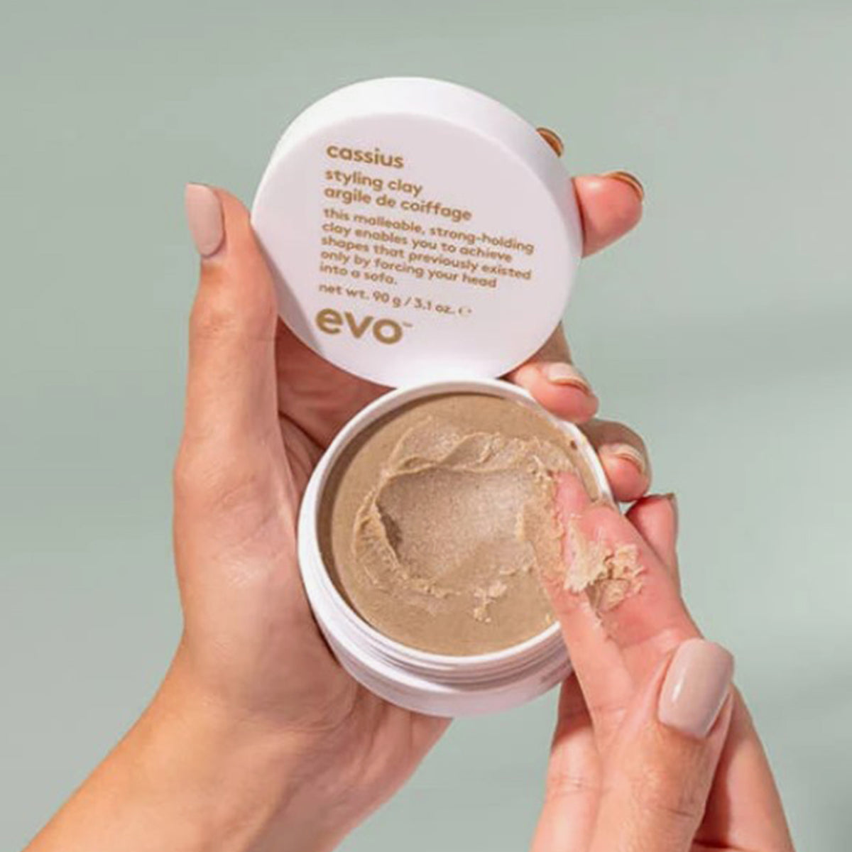 Hände halten einen offenen Tiegel Evo Cassius Styling Clay, mit einer kleinen Menge des Produkts auf zwei Fingerspitzen.
