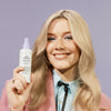 Lächelnde Frau mit langen blonden Haaren hält eine Flasche Fabuloso Platinum Blonde Toning Mist.