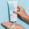 Vor einem blauen Hintergrund drückt eine Person weiße Lotion aus einer Tube Head Mistress Cuticle Sealer auf ihre Handfläche.