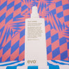 Eine weiße Flasche Icon Welder Hitzeschutzspray vor einem rot-blauen geometrischen Hintergrund.