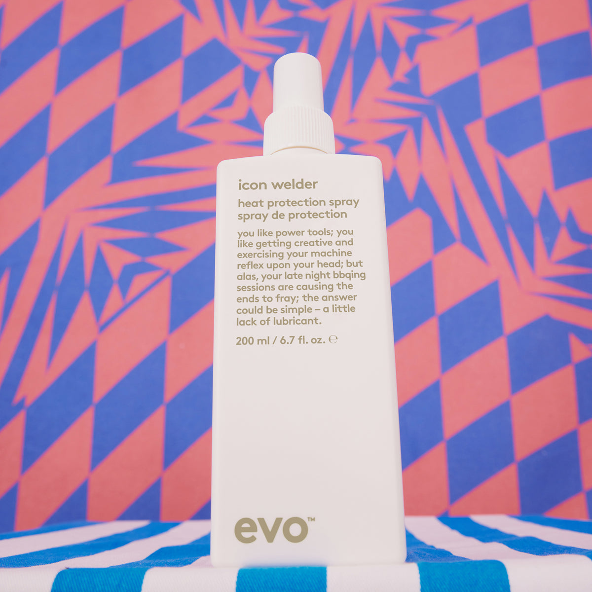 Eine weiße Flasche Icon Welder Hitzeschutzspray vor einem rot-blauen geometrischen Hintergrund.