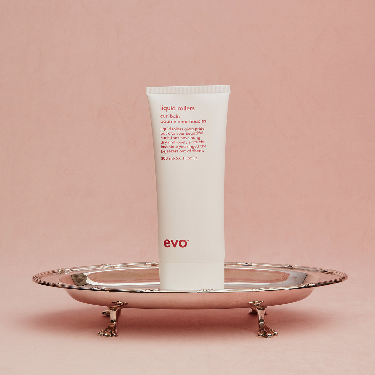 Eine weiße Tube Liquid Rollers Curl Balm steht aufrecht auf einem silbernen Tablett vor einem rosa Hintergrund.