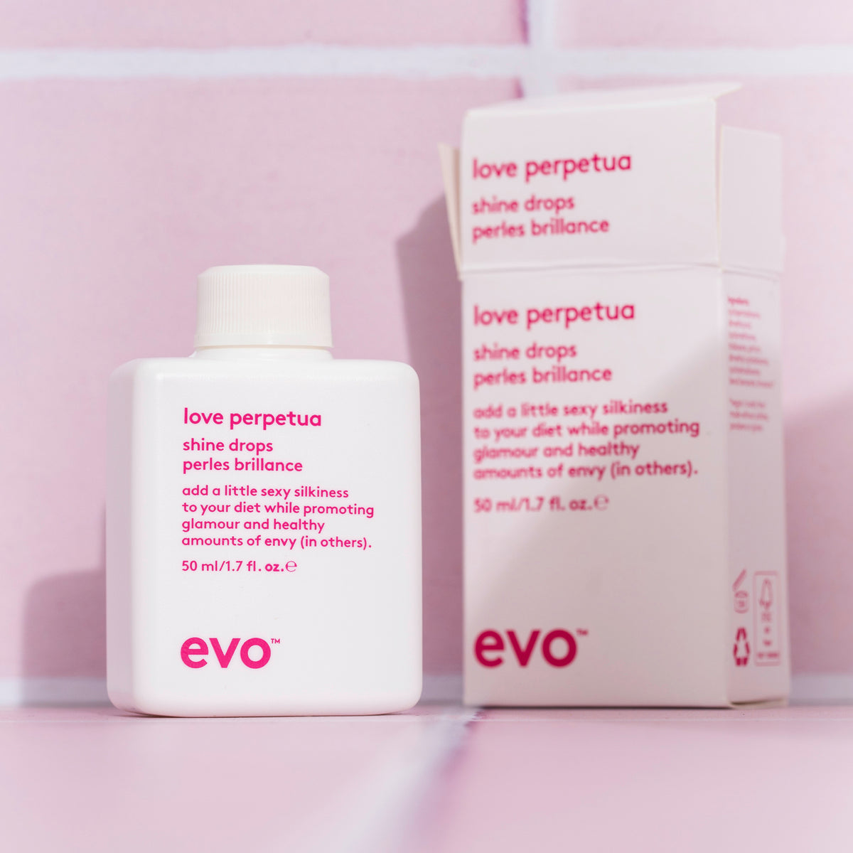 Weiße Flasche und eine Schachtel Love Perpetua Shine Drops auf einem rosa gekachelten Hintergrund präsentiert.