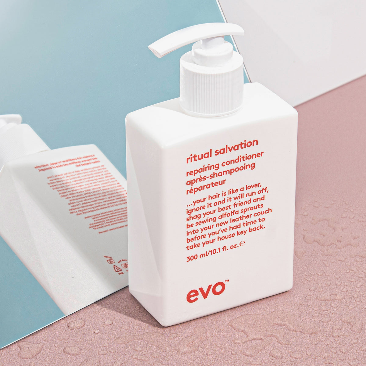 Weiße Pumpflasche Ritual Salvation Repairing Conditioner steht auf einer rosa Oberfläche neben einem Spiegel, umgeben von Wassertropfen.